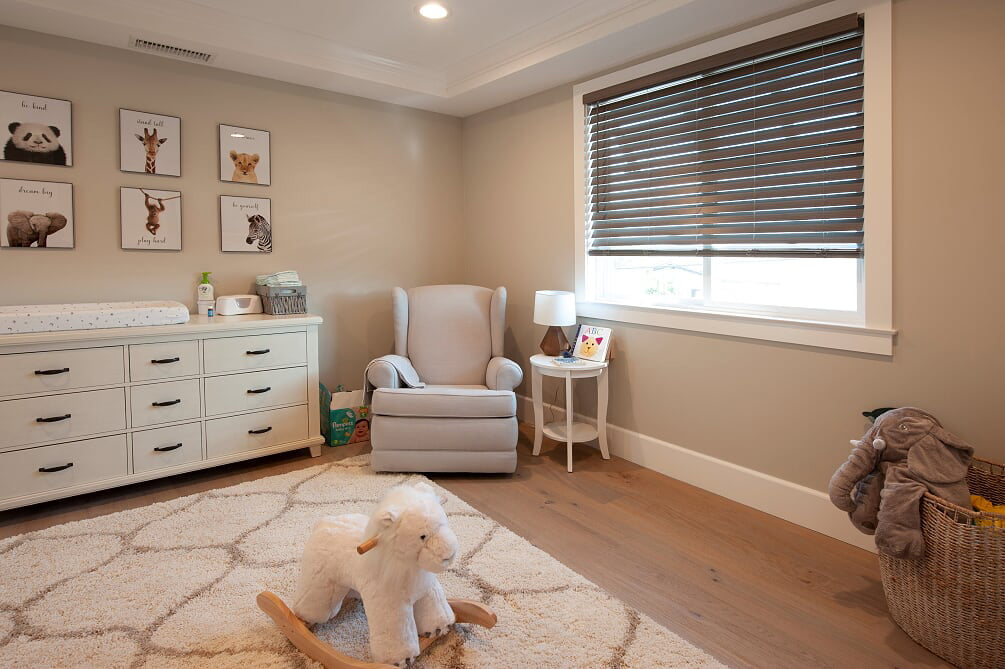 A cozy nursery decorated with soft, neutral tones. Norman's SmartPrivacy Blinds cover the window, allowing adjustable light control. The room features a comfortable rocking chair beside a small side table with a lamp and a children's book. A dresser with a changing pad sits against the wall, adorned with animal-themed artwork above. A plush rug and a rocking sheep toy on the floor add warmth and charm to the space, creating an inviting environment for a little one.