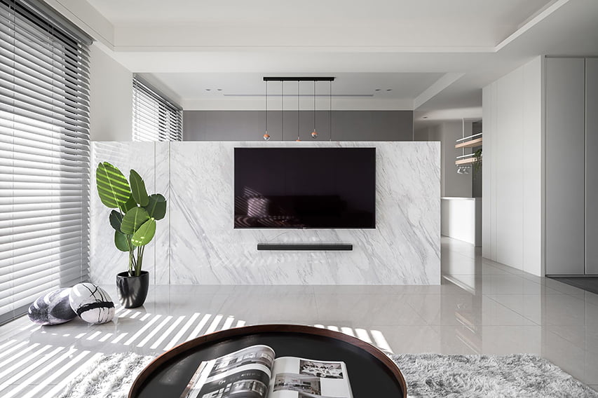 A modern living room with a marble accent wall that holds a flat-screen TV and a sleek soundbar. Norman's SmartPrivacy Blinds cover the large windows, allowing filtered natural light to create subtle patterns across the floor. A potted plant and decorative stone accents add a touch of nature to the clean, minimalist space. The room includes a shaggy rug and a round coffee table with an open magazine, enhancing the cozy yet sophisticated ambience.