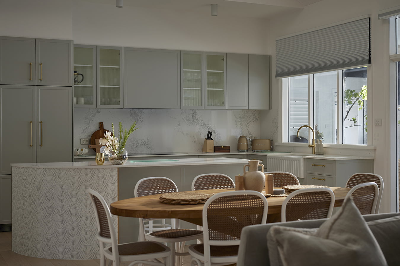 Stylish kitchen with soft gray cabinetry, brass hardware, and a marble-patterned backsplash. A mosaic-tiled kitchen island is decorated with a vase of flowers, adding a touch of elegance. The large window above the sink is fitted with Norman Honeycomb Shades, allowing soft natural light into the room. The dining area features a wooden table and woven-back chairs, enhancing the room’s warm, inviting atmosphere.