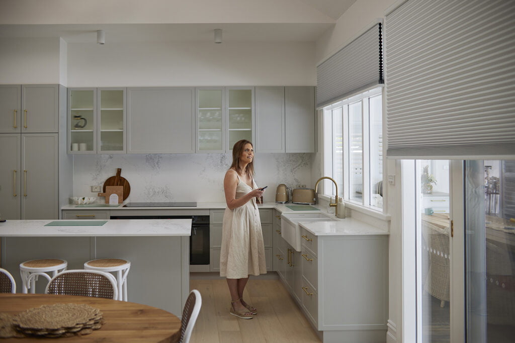 A modern kitchen with grey cabinetry and gold accents features motorised Honeycomb Shades on the windows, offering seamless light control and privacy. A woman stands by the counter, holding a remote to adjust the shades effortlessly