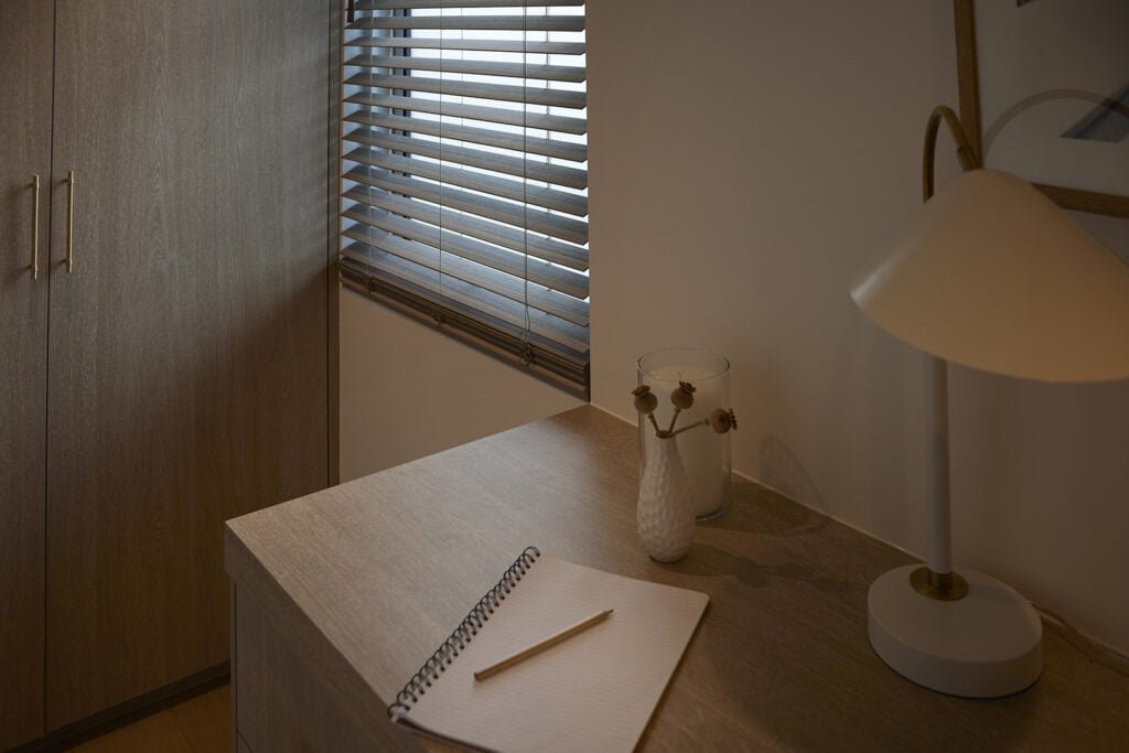 A cozy workspace illuminated by soft lighting, featuring Norman's SmartPrivacy Blinds in a light wood finish. The setting includes a minimalist desk with a notebook, pencil, and a vase with dried flowers, alongside a contemporary desk lamp. The SmartPrivacy Blinds provide both style and privacy, enhancing the calm and focused atmosphere of the room.