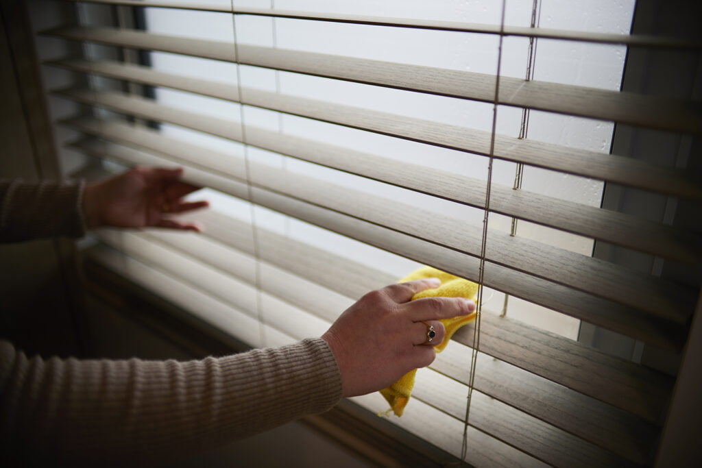 Close-up of hands gently cleaning Norman's SmartPrivacy Blinds with a yellow cloth. The blinds feature a light wood texture, offering both style and functionality with easy maintenance. The person’s hands carefully wipe along the slats, showcasing the durability and user-friendliness of the blinds.