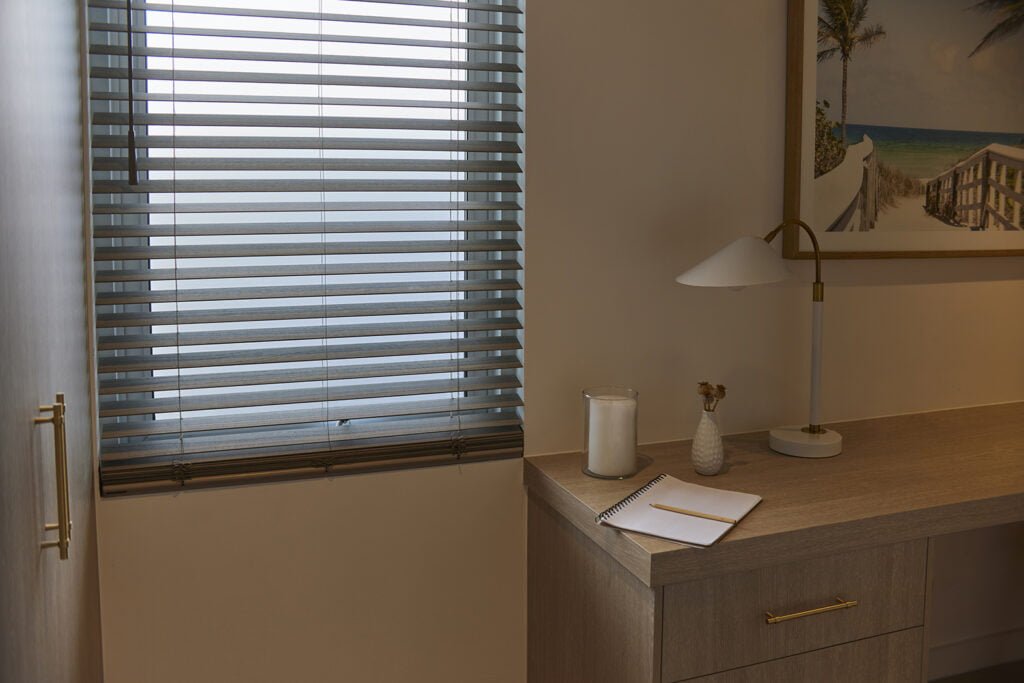 A cozy home office nook featuring Norman's SmartPrivacy Blinds, offering enhanced privacy and light control. The soft, muted light filtering through the blinds creates a calm ambiance. A wooden desk holds a minimalist table lamp, a notebook, a candle, and a small decorative vase, while a framed coastal photograph adds a touch of warmth to the space.