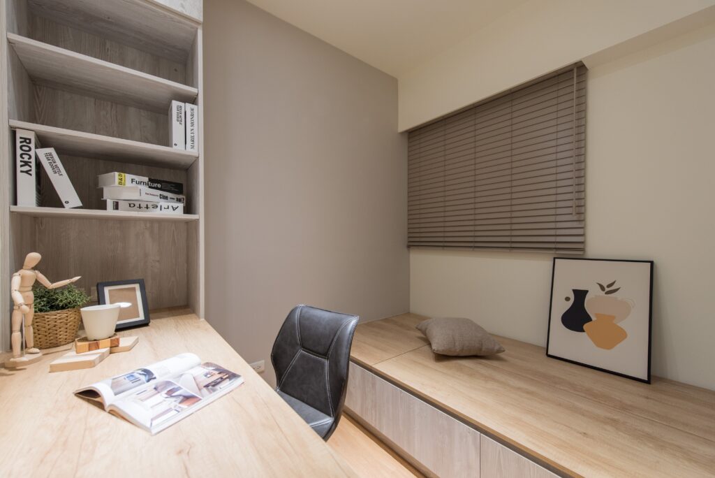 A modern and minimalist study area featuring Norman’s FauxWood Blinds in a warm taupe colour. The room is styled with a natural wood desk, decorated with art books, a small potted plant, and a figurine. The cozy built-in bench below the blinds adds seating, complemented by a cushion and framed artwork. This setup combines functionality with a soft, warm aesthetic, perfect for focused work and relaxation.