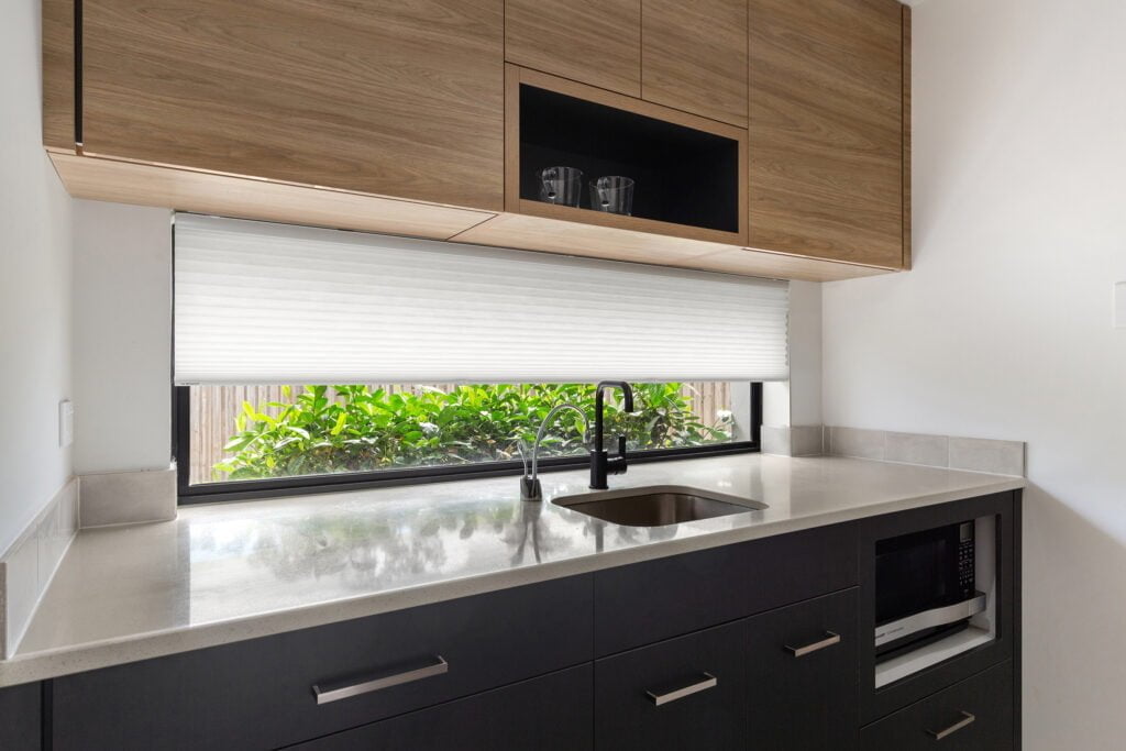 A sleek, modern kitchen with dark cabinetry and a smooth, light countertop. Above the countertop, there’s a large window covered with Norman's Honeycomb Shades, partially drawn to allow soft light while maintaining privacy. The black faucet and sink contrast with the bright surroundings, and natural greenery outside enhances the fresh, inviting feel of the space. Upper cabinets in wood add warmth, creating a balanced, contemporary design.