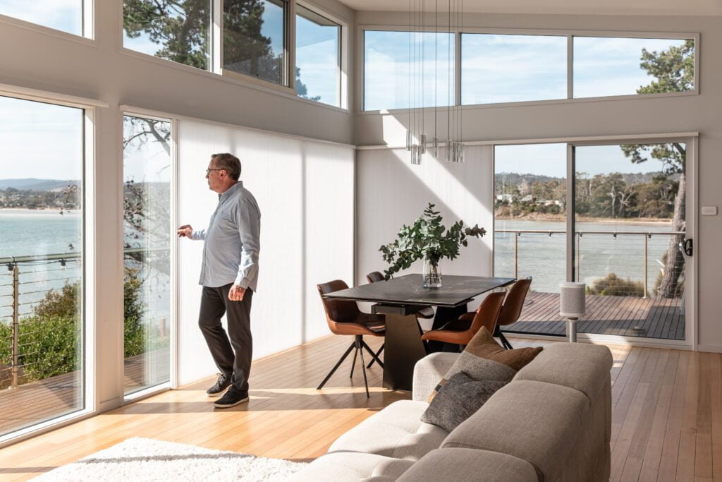 A spacious, modern living and dining area with floor-to-ceiling windows overlooking a scenic water view. The room is filled with natural light, and Norman's Patio Door Vertical Honeycomb Shades are partially drawn to control the sunlight while maintaining the airy feel. A man stands by the window, appreciating the view. The dining area features a round table with leather chairs and a vase of greenery, while the adjacent living space has a cozy, light-coloured sofa. The open layout and expansive windows emphasise the connection between the indoor space and the outdoor deck, creating a serene and welcoming atmosphere.