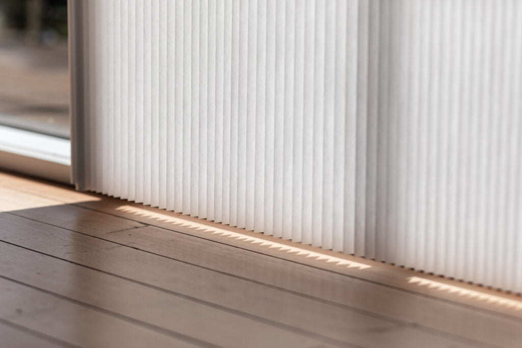 A close-up of Norman's Patio Door Vertical Honeycomb Shades in a bright room. The shades softly diffuse the sunlight, casting a subtle shadow pattern onto the hardwood floor, highlighting the shades' texture and light-filtering properties.