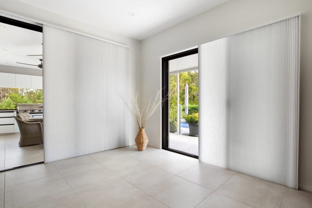 A modern, minimalistic room with large sliding glass doors leading to an outdoor patio. The doors are covered with Norman's Patio Door Vertical Honeycomb Shades, which are fully extended to provide privacy and light control. The shades have a soft, textured appearance, adding an elegant touch to the space. Natural light filters through gently, maintaining a bright ambiance while shielding the room from direct sunlight. A decorative wicker vase with long branches stands in the corner, enhancing the room's organic and airy feel.