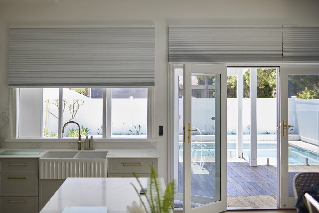 A bright, modern kitchen with large windows and doors opening to an outdoor pool area. The windows are covered with Norman's Honeycomb Shades, which are pulled down to provide privacy and light control. Below the window is a farmhouse-style sink with a brass faucet, set against a white countertop and light cabinetry with brass handles. French doors on the right lead outside, allowing for a seamless indoor-outdoor flow, enhancing the open, airy feel of the space.