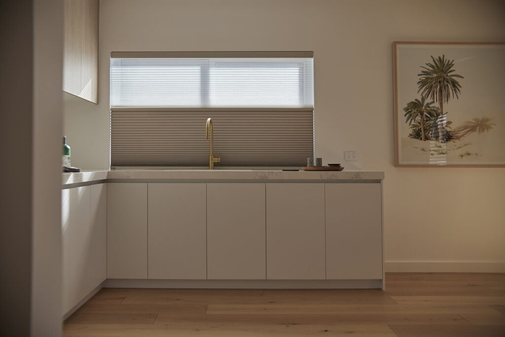 Modern kitchen with minimalist design, featuring white cabinetry and a sleek marble countertop. A gold faucet stands out against the neutral tones, positioned beneath a window fitted with Norman's Day and Night Honeycomb Shades, which filter soft natural light through. A framed artwork of palm trees adds a touch of warmth and character to the space.