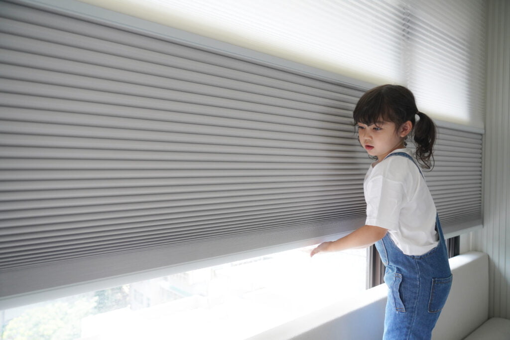 A young girl dressed in denim overalls stands by a large window, adjusting Norman's Honeycomb Shades. The child-friendly shades are easy to operate, providing a balance of light control and privacy while showcasing a safe, cordless design. The background view includes a cityscape with greenery and buildings, highlighting the versatile application of the shades in urban environments.