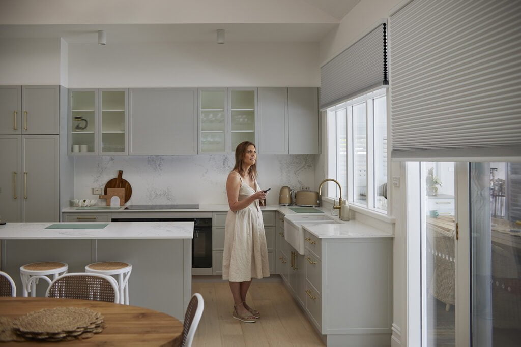 A modern kitchen featuring Norman's Honeycomb Shades with motorisation. The shades are partially lowered on a large window, allowing filtered light to enter the space. A woman stands near the counter, holding a remote control, presumably adjusting the shades. The kitchen has soft gray cabinetry with brass hardware, a farmhouse sink, and a brass faucet, complementing the neutral tones and sleek design. A round wooden dining table with woven chairs sits in the foreground, adding a touch of warmth and texture to the room.
