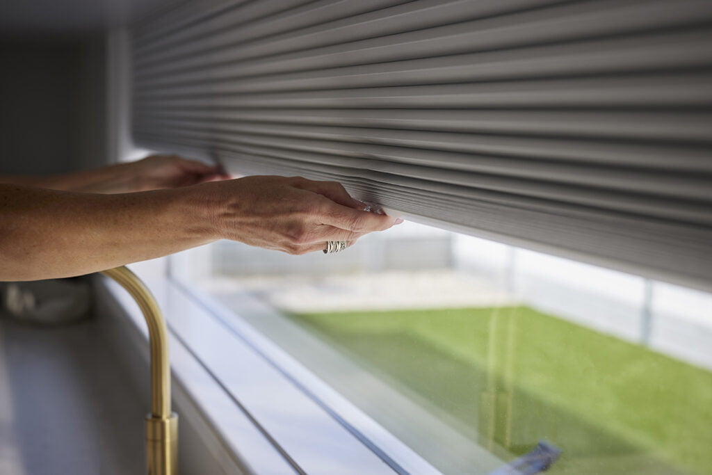 Close-up of hands adjusting Norman's Honeycomb Shades, demonstrating the shade's smooth, easy-to-lift operation. The window in the background overlooks a green outdoor space, adding a touch of natural light to the scene.