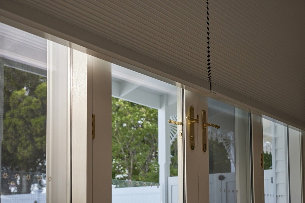 Close-up of Norman's motorised Honeycomb Shades covering a large window or door with brass handles. The shades offer privacy while still allowing a view of the greenery outside, showcasing both functionality and elegance in window treatment.