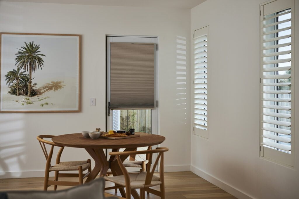 Bright dining nook featuring a round wooden table with matching chairs, set against a wall adorned with a framed artwork of palm trees. The room includes a door with Norman Honeycomb Shades partially drawn, alongside tall windows with Norman's Woodlore Shutters that allow natural light to filter through, casting soft shadows on the walls. The space combines natural textures with a serene, minimalist aesthetic.