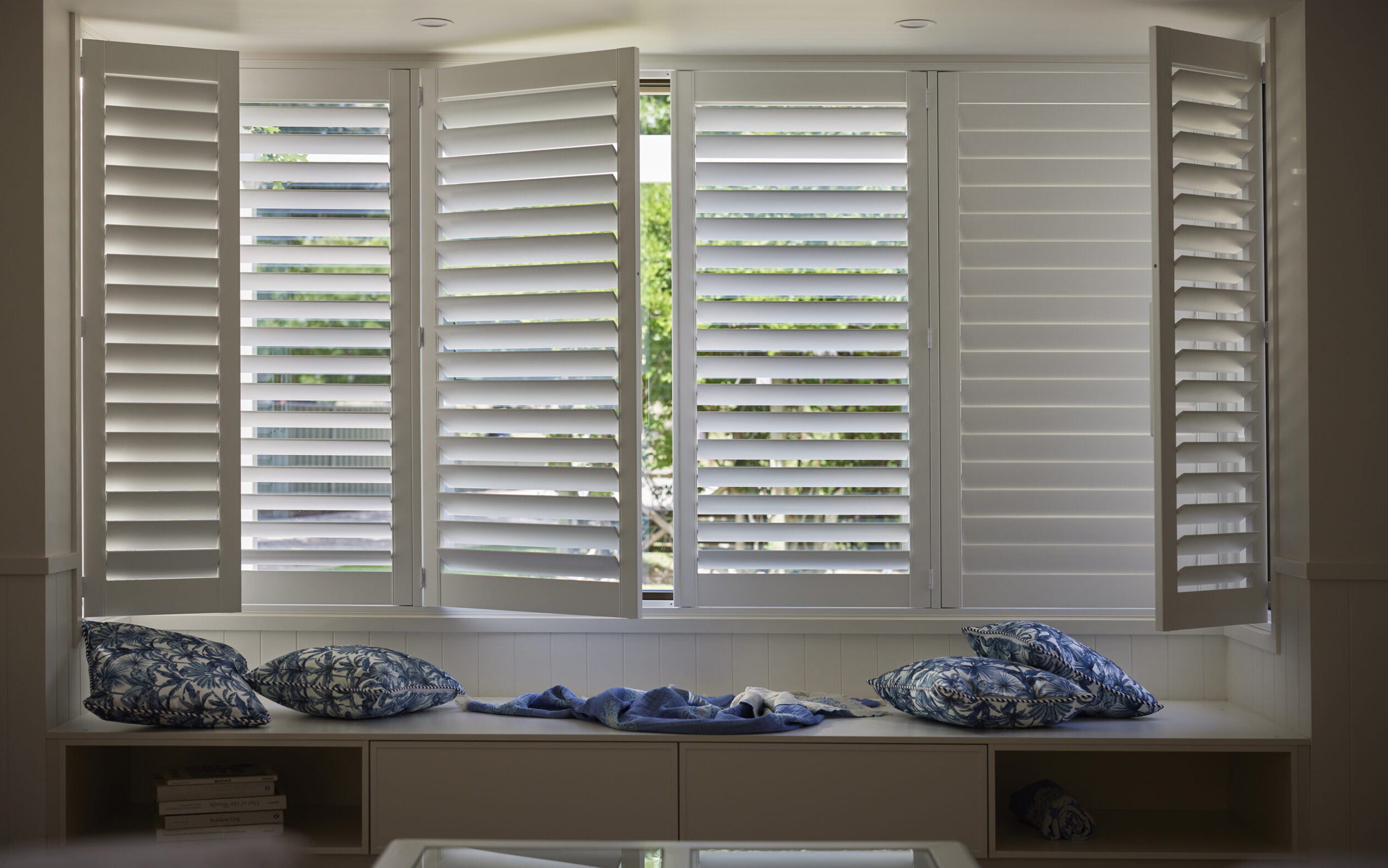 Wide-angle view of a cozy window seat framed by Norman Woodlore Shutters, showcasing their clean lines and functional design, allowing adjustable natural light and privacy.