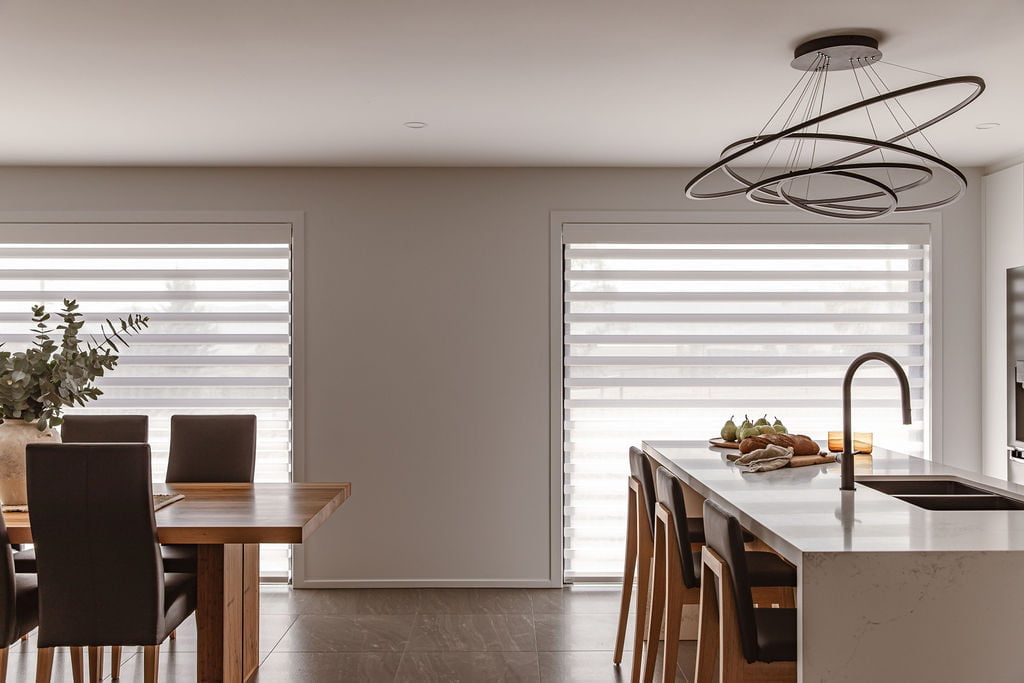 A modern dining and kitchen area with Norman's PerfectSheer Shades on two large windows. The shades offer a balance of privacy and natural light, creating a warm and inviting atmosphere. The space features a dining table with dark chairs, a kitchen island with seating, and a stylish, circular pendant light above.