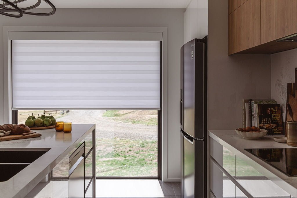 A modern kitchen with Norman's PerfectSheer Shades on a large window, filtering natural light while maintaining a level of privacy. The soft, layered fabric design complements the minimalist decor, featuring clean lines, a sleek refrigerator, and a countertop with fresh produce, creating a warm, inviting atmosphere.