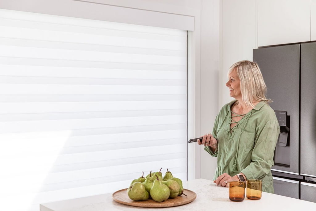 A woman in a modern kitchen using a remote control to adjust Norman's PerfectSheer Shades. The shades are elegantly designed, providing both privacy and light control while blending seamlessly into the contemporary decor. On the kitchen counter, there is a plate of green pears and two amber glasses, adding a touch of colour to the neutral space.