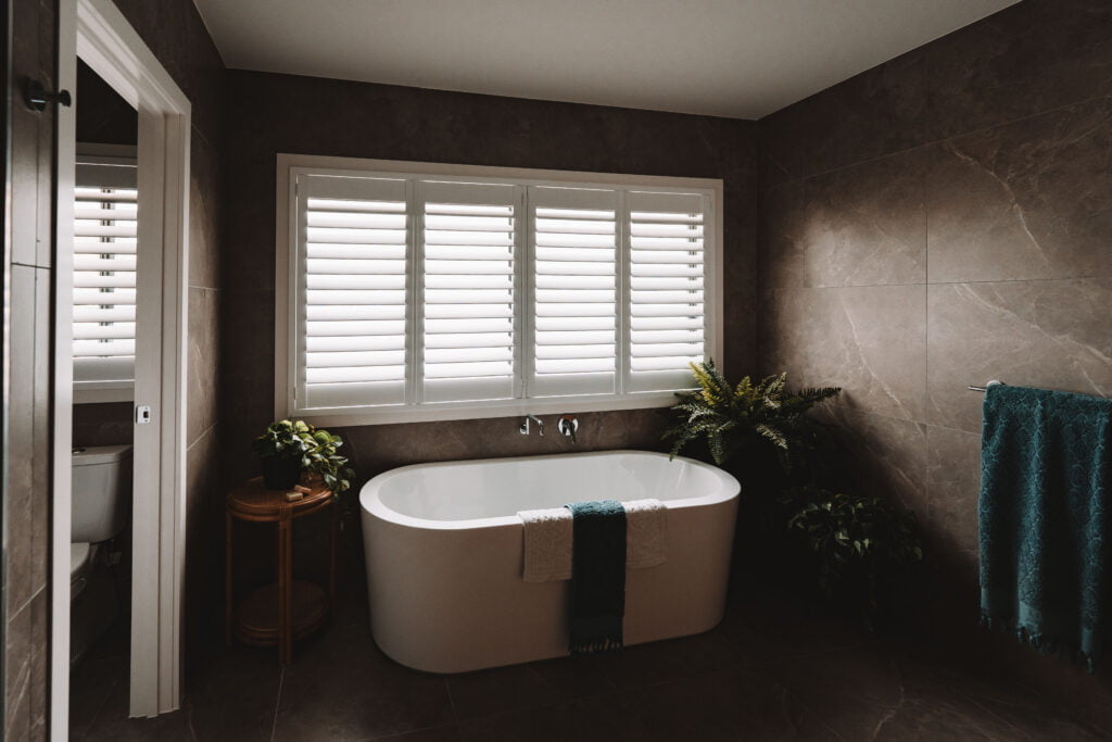 Bathroom featuring Norman Shutters on a large window above a freestanding bathtub, surrounded by modern dark tiles and greenery, showcasing a stylish and practical window covering solution.