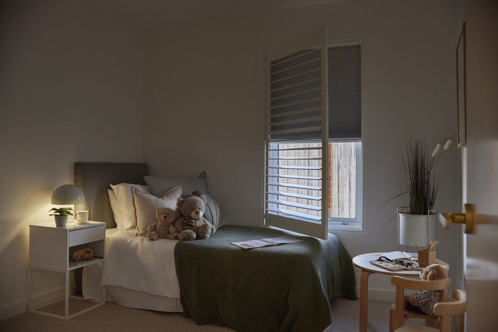 Cozy child’s bedroom featuring Norman’s Blockout Shutter for optimal light control and privacy. The warm ambiance highlights a neatly made bed with plush teddy bears, a soft green blanket, and a bedside table illuminated by a soft lamp.