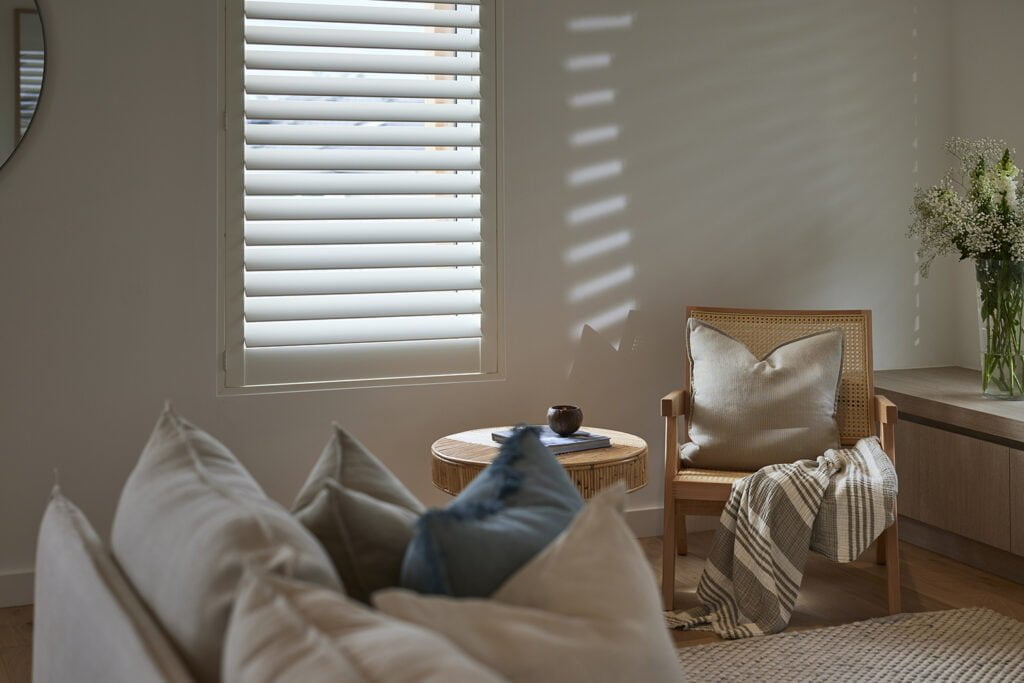 Serene living space featuring Norman’s Shutters, casting soft light patterns across the room. Styled with cozy neutral tones, a rattan accent chair, plush pillows, and a textured throw, this space exudes calm and understated elegance.