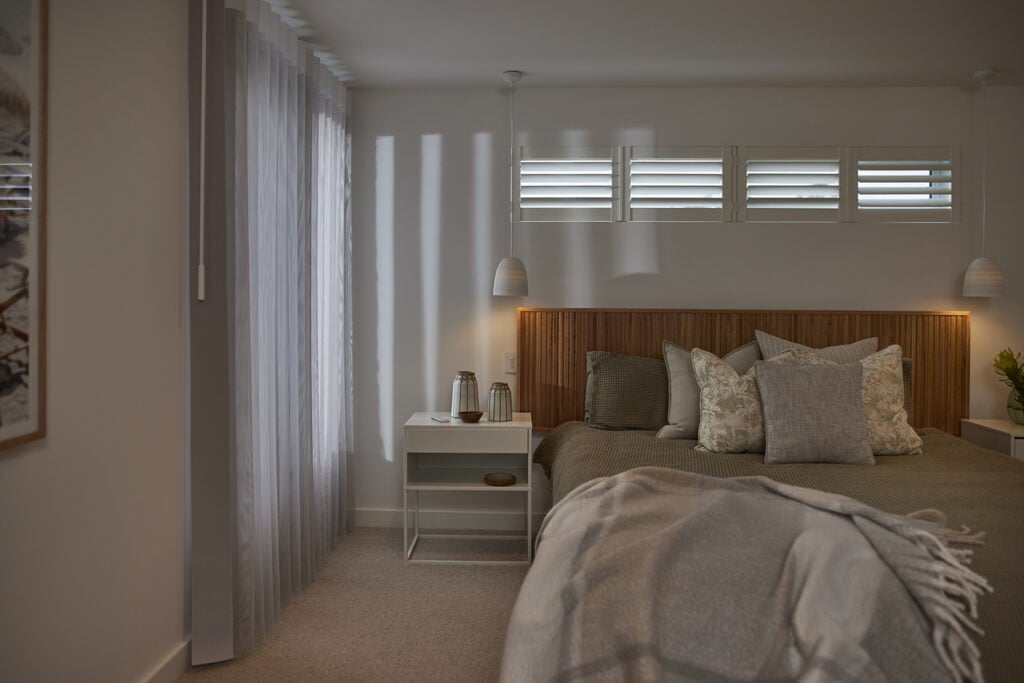 Cozy bedroom with a modern aesthetic featuring a wooden headboard, layered pillows, and a soft blanket. Vertical sheer curtains allow soft, diffused light to enter, complemented by small, horizontal windows with Norman Woodlore Shutters above the bed. The bedside tables are styled with decorative lanterns and a minimal pendant light hangs on each side, creating a warm and inviting atmosphere.