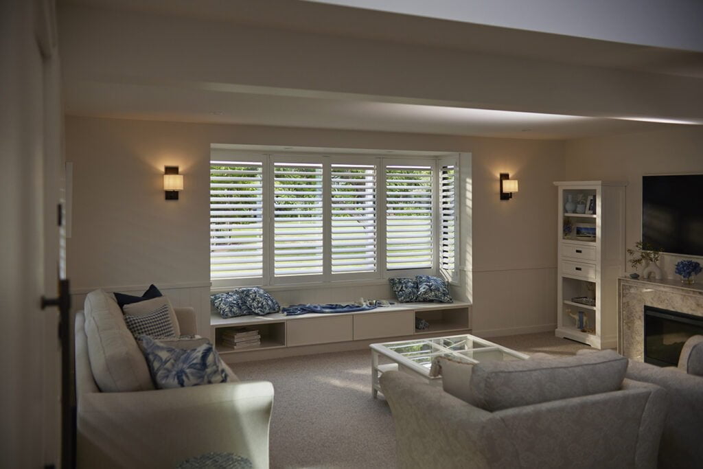 Elegant living room featuring Norman’s Shutters on a large bay window, framing a tranquil view of lush greenery. The space is styled with cozy seating, soft neutral tones, and a charming window seat adorned with blue-patterned cushions, creating a warm and inviting atmosphere.
