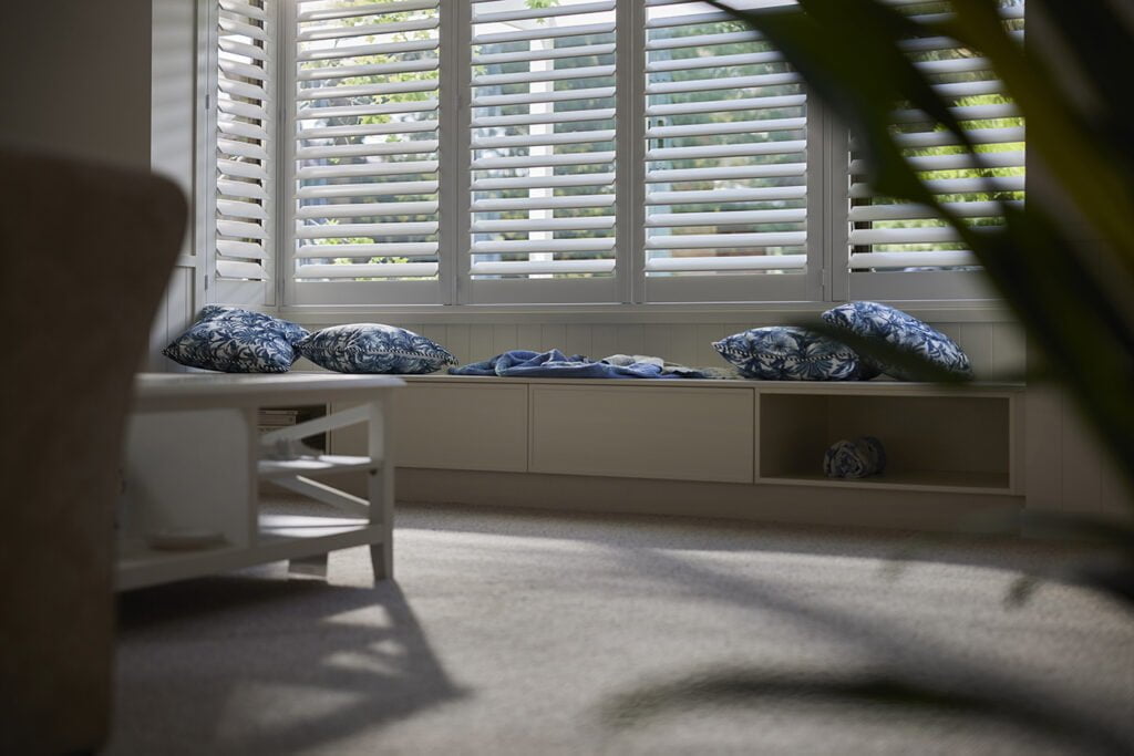 Close-up of a cozy window seat framed by Norman’s Shutters, allowing natural light to filter through while providing privacy. Styled with blue patterned cushions and a soft throw, this nook is perfect for relaxing and enjoying the view.