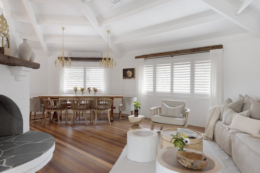 A bright and airy dining and living space featuring Norman’s Shutters on the windows. The room has vaulted ceilings painted white with exposed wooden beams. The dining area includes a long wooden table with multiple chairs, and an elegant chandelier hangs above. The living area is styled with a large beige sofa, a couple of wooden coffee tables, and decorative plants, creating a cozy, inviting atmosphere.