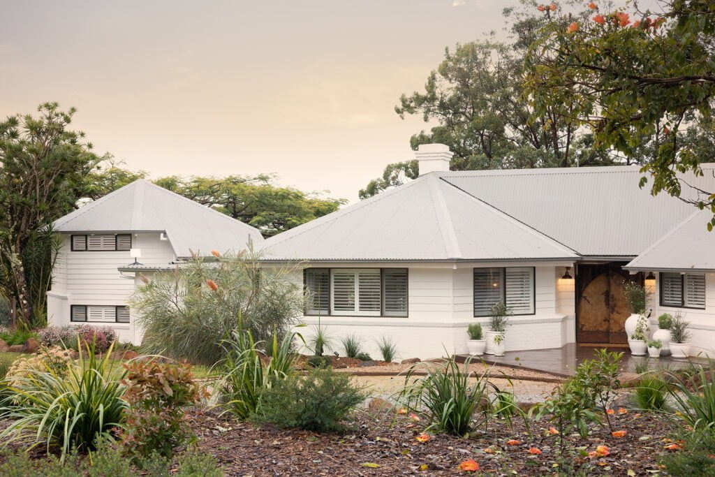 Beautiful exterior view of a charming home featuring Norman’s Shutters, adding a touch of elegance and functionality to the windows. Surrounded by lush greenery and a thoughtfully landscaped garden, the home exudes a serene and welcoming vibe.