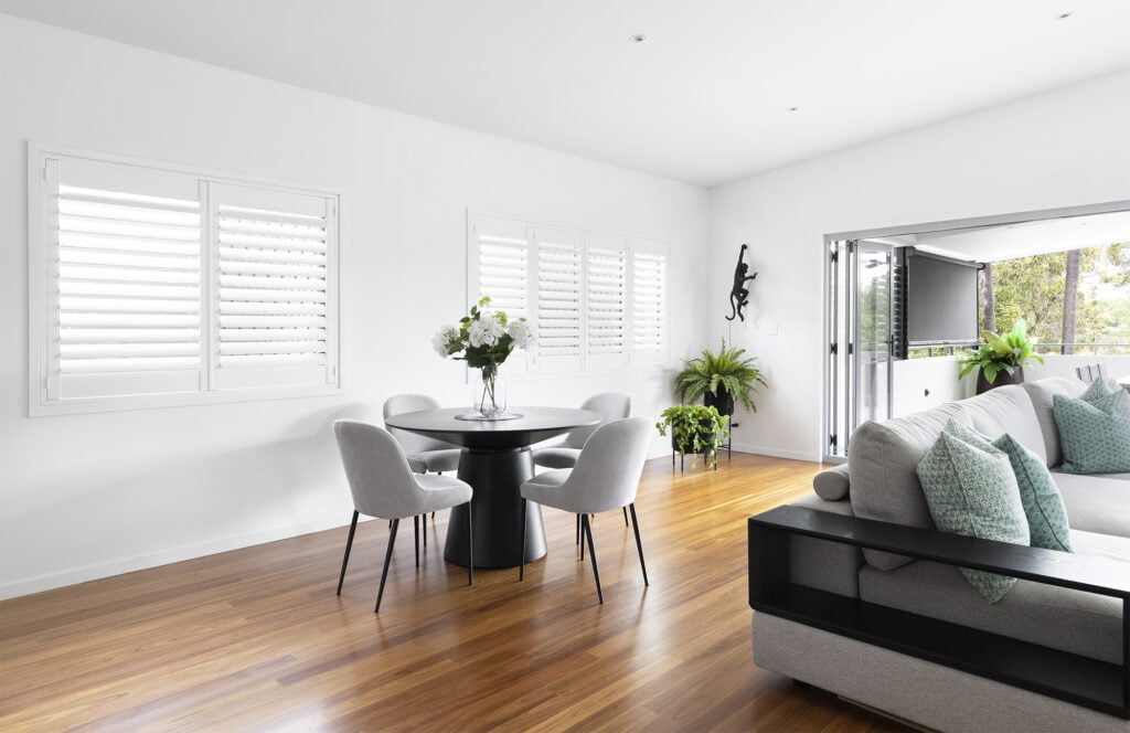 Modern living and dining space featuring Norman Shutters on large windows, complementing the bright, open design with natural light. The room showcases a stylish round dining table, grey upholstered chairs, and greenery, creating a fresh and inviting atmosphere.