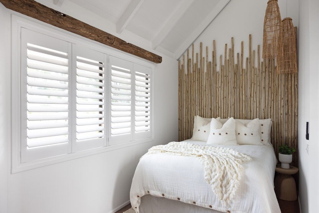 Bright and airy bedroom featuring Norman’s Brightwood Shutters, offering light control and privacy. The space combines rustic charm with modern elegance, highlighted by natural wood accents, woven pendant lights, and a cozy, textured bed dressed in soft neutrals.