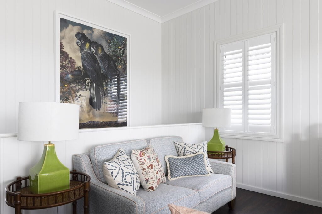 Bright and cozy living room featuring Norman’s Shutters, allowing soft natural light to filter through. The space is styled with a patterned sofa, vibrant throw pillows, bold green lamps, and a striking black cockatoo artwork, creating a sophisticated yet inviting atmosphere.