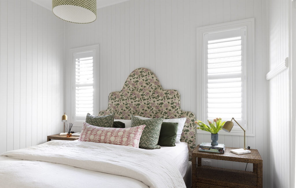Cozy bedroom featuring Norman Shutters on tall windows, allowing soft natural light to filter through. The space is styled with a floral upholstered headboard, layered cushions, and wicker bedside tables, creating a charming and inviting retreat