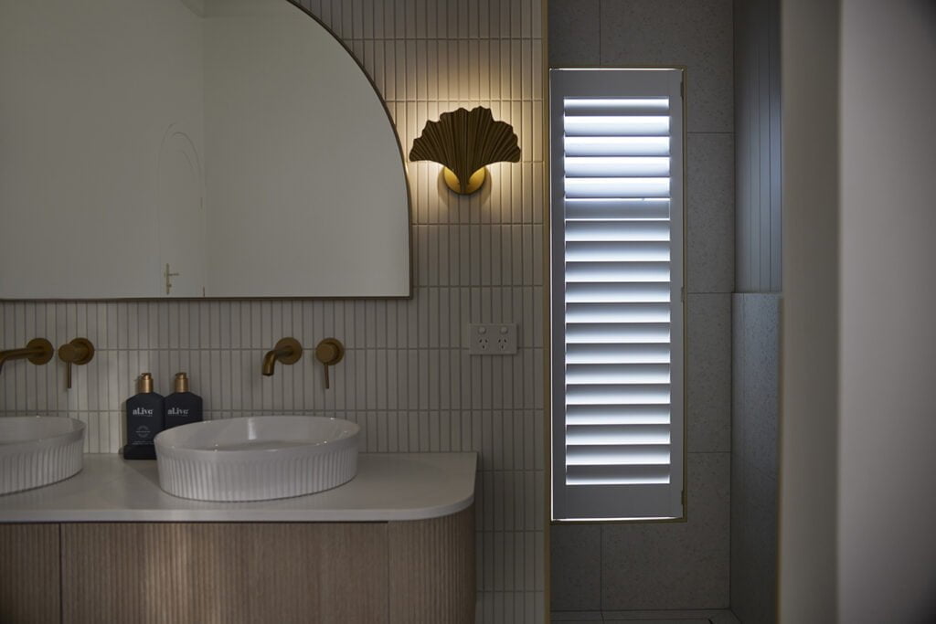 Elegant bathroom featuring Norman’s Woodlore Plus Waterproof Shutters, providing light control and privacy while standing up to moisture. Styled with warm brass accents, a curved mirror, and soft ambient lighting, this space combines luxury with practicality.
