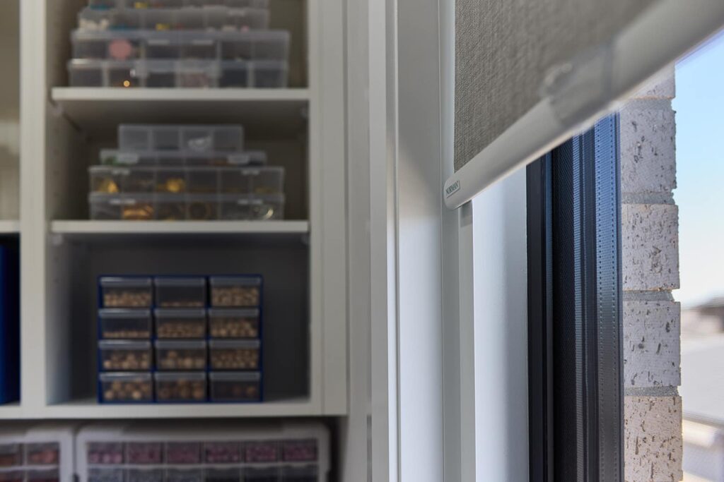 Close-up view of Norman’s Soluna Roller Shades with LightGuard installed in a window, displaying their precision fit and light-blocking edges. The shades are partially drawn, revealing a glimpse of the cityscape outside. In the foreground, a well-organized craft storage system holds various art supplies, highlighting the functional use of space in a creative workroom.