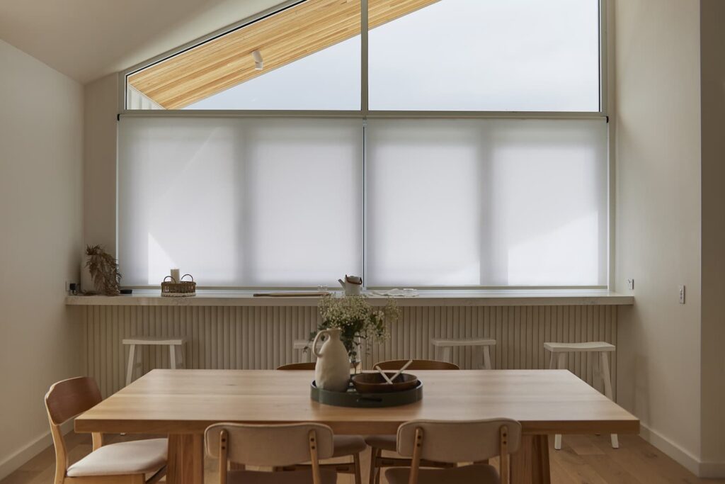 A minimalist dining area bathed in soft, diffused light from Norman’s Soluna Roller Shades covering large windows. The room features a simple wooden dining table surrounded by matching chairs, a quaint bench by the wall, and decorative elements such as a ceramic rabbit vase and dried plants. The tranquil setting is ideal for serene dining experiences, enhancing the space with both style and functionality
