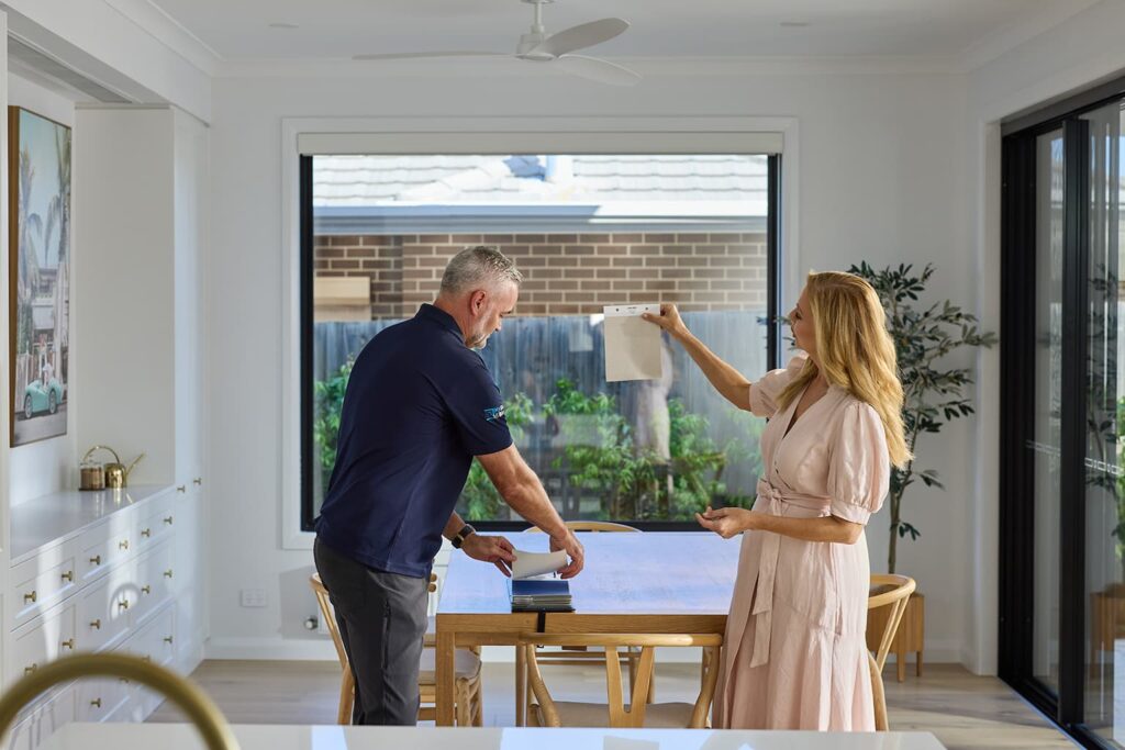 In a modern, brightly lit kitchen, a woman in a pink dress holds up a fabric sample of Soluna Roller Shades for a man in a navy blue uniform to examine. They stand over a wooden dining table covered with various shade samples and tools, discussing options. The scene includes a sleek white kitchen counter and large glass doors that lead to a garden, showcasing the indoor-outdoor flow of the home.