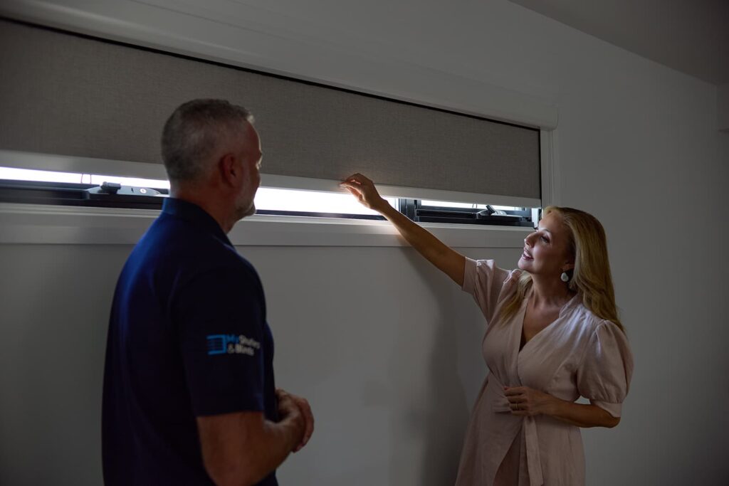 A woman in a pink dress demonstrates the effectiveness of Norman’s Soluna Roller Shades with LightGuard 360 to a man in a dark blue uniform. They are in a dimly lit room, with the shade partially drawn to showcase the light filtering technology. The woman points towards the slender gap at the top of the shade, highlighting the minimal light leakage, which underscores the shade’s superior design for light control and privacy.