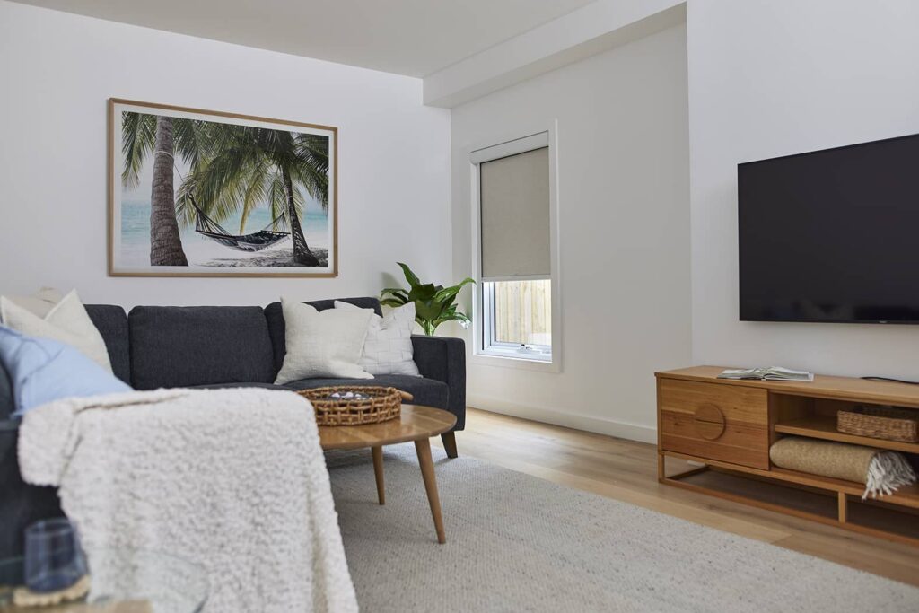 A cozy living room featuring Norman’s Soluna Roller Shades with LightGuard 360 on the windows, providing effective light control and privacy. The room is stylishly decorated with a dark grey sofa adorned with white cushions, a textured white throw, a tropical beach scene framed on the wall, and a wooden TV stand. Soft natural light filters through, creating a warm, inviting atmosphere.