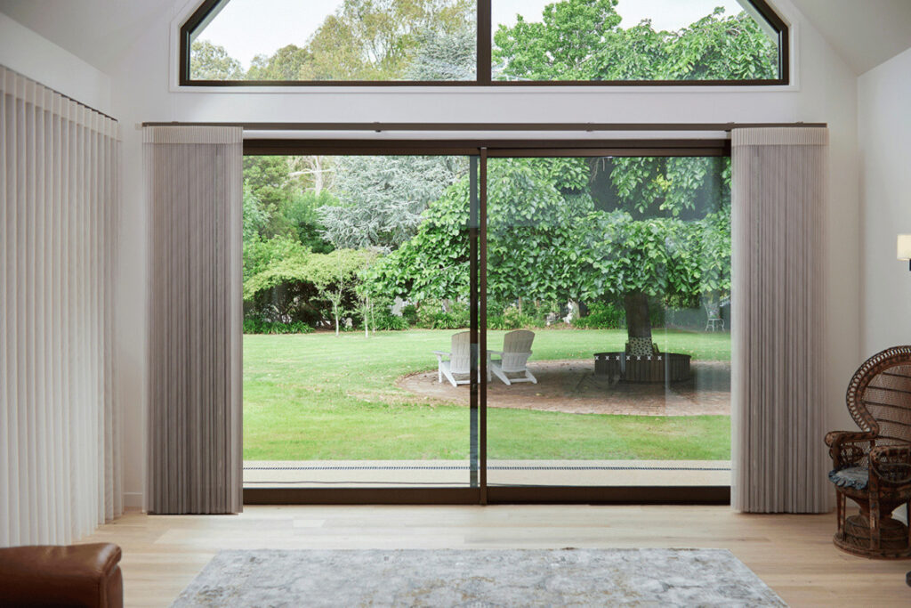 A bright living room with large sliding glass doors, showcasing Norman’s SmartDrape Shades drawn to the sides to reveal a verdant garden. The room is elegantly designed with a contemporary style, featuring a leather armchair, a patterned rug, and a classic wicker chair beside a fireplace. The peaceful outdoor view includes two white Adirondack chairs under a tree, enhancing the tranquil atmosphere of the space.