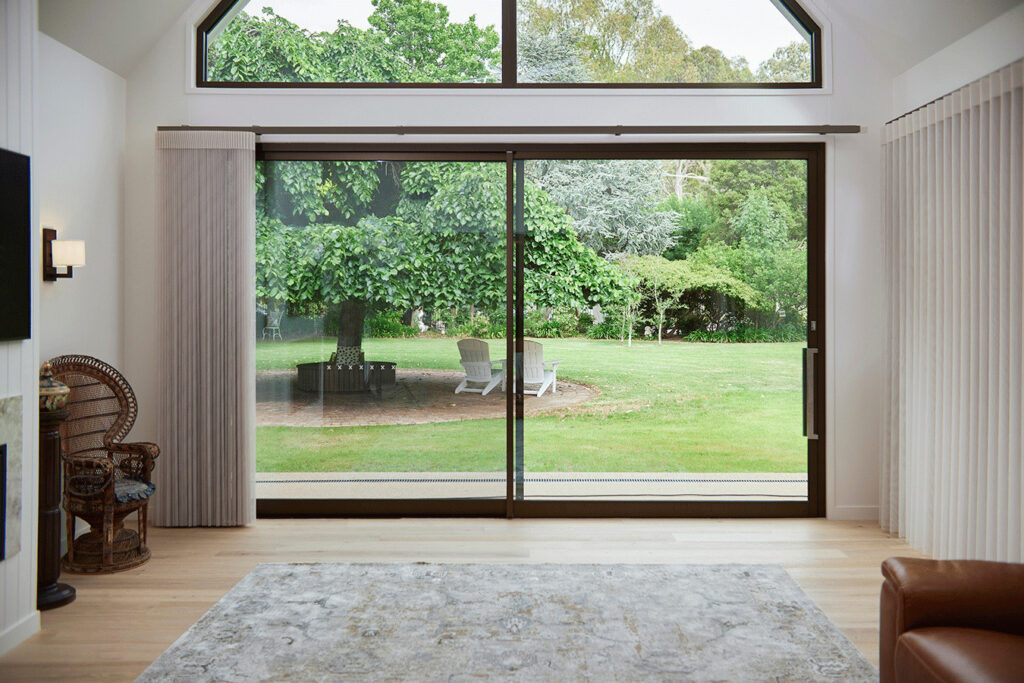 A modern living room with a large sliding glass door framed by Norman’s SmartDrape Shades, offering a picturesque view of a lush garden with two Adirondack chairs under a large tree. The interior is elegantly designed with a light wood floor, a patterned rug, a brown leather armchair, and a vintage wicker chair by a fireplace, creating a tranquil and inviting environment.