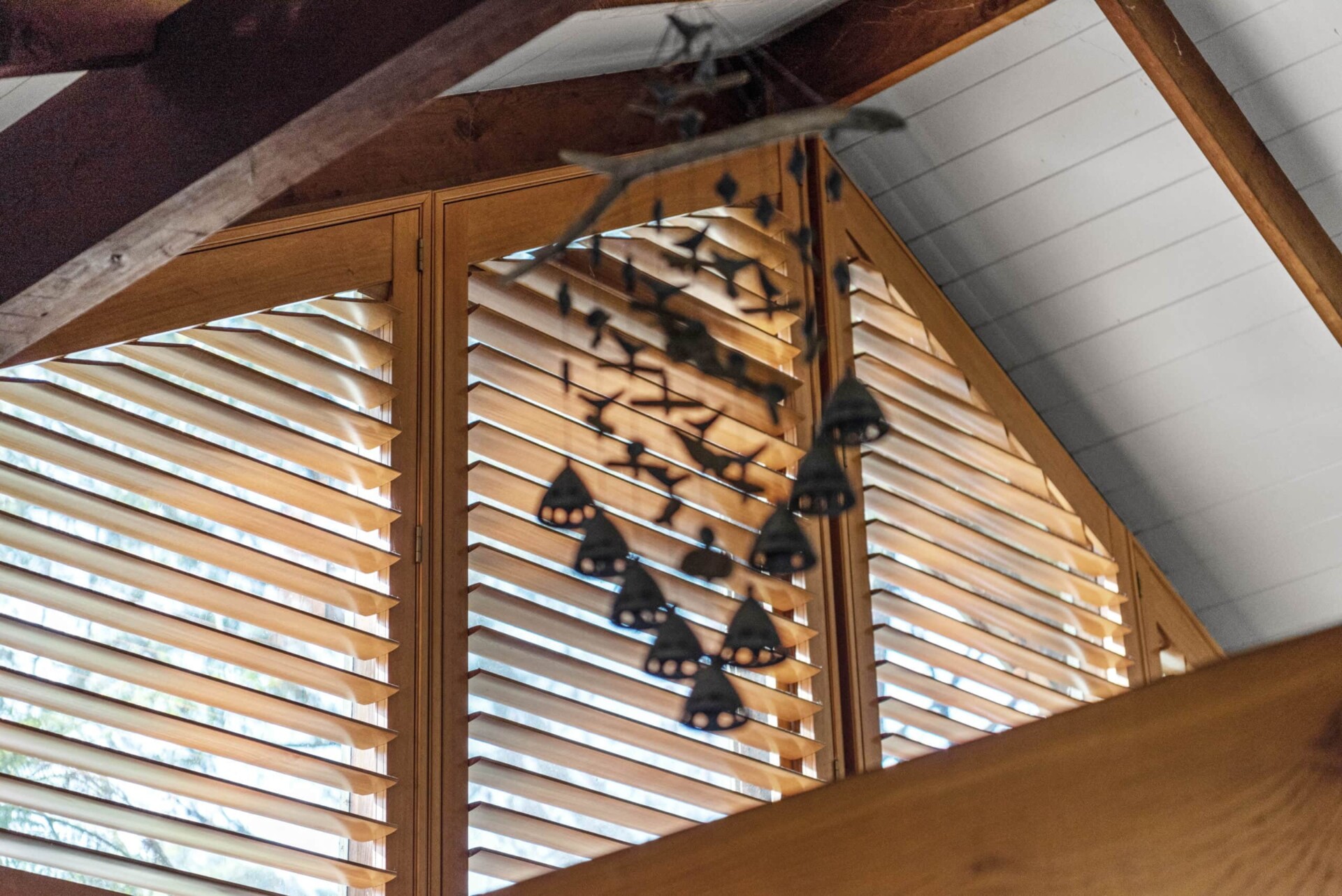 Warm-toned wooden shutters from the Normandy range fitted on a triangular window in a high-ceilinged space. The shutters allow natural light to filter through while complementing the exposed timber beams of the ceiling. A decorative hanging mobile in the foreground adds a touch of rustic charm to the serene and inviting atmosphere.