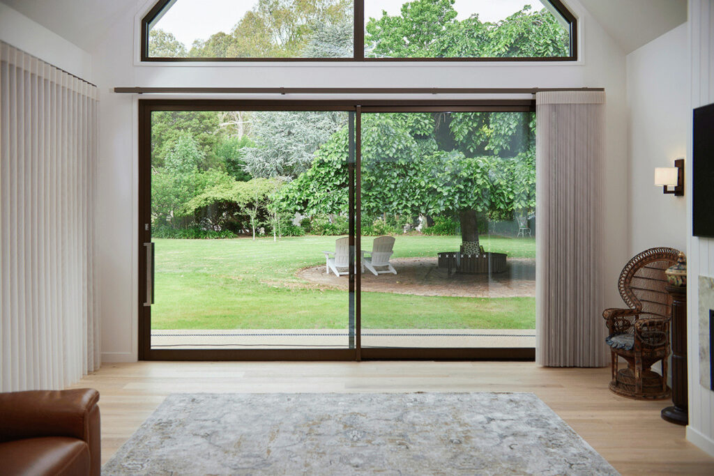 A luxurious living room with Norman’s SmartDrape Shades framing a large glass sliding door that opens up to a lush garden view. The shades are drawn to the sides, letting in ample natural light that highlights the modern interior, including a brown leather armchair, a patterned rug, and a wicker chair by the window, creating a tranquil and inviting space.